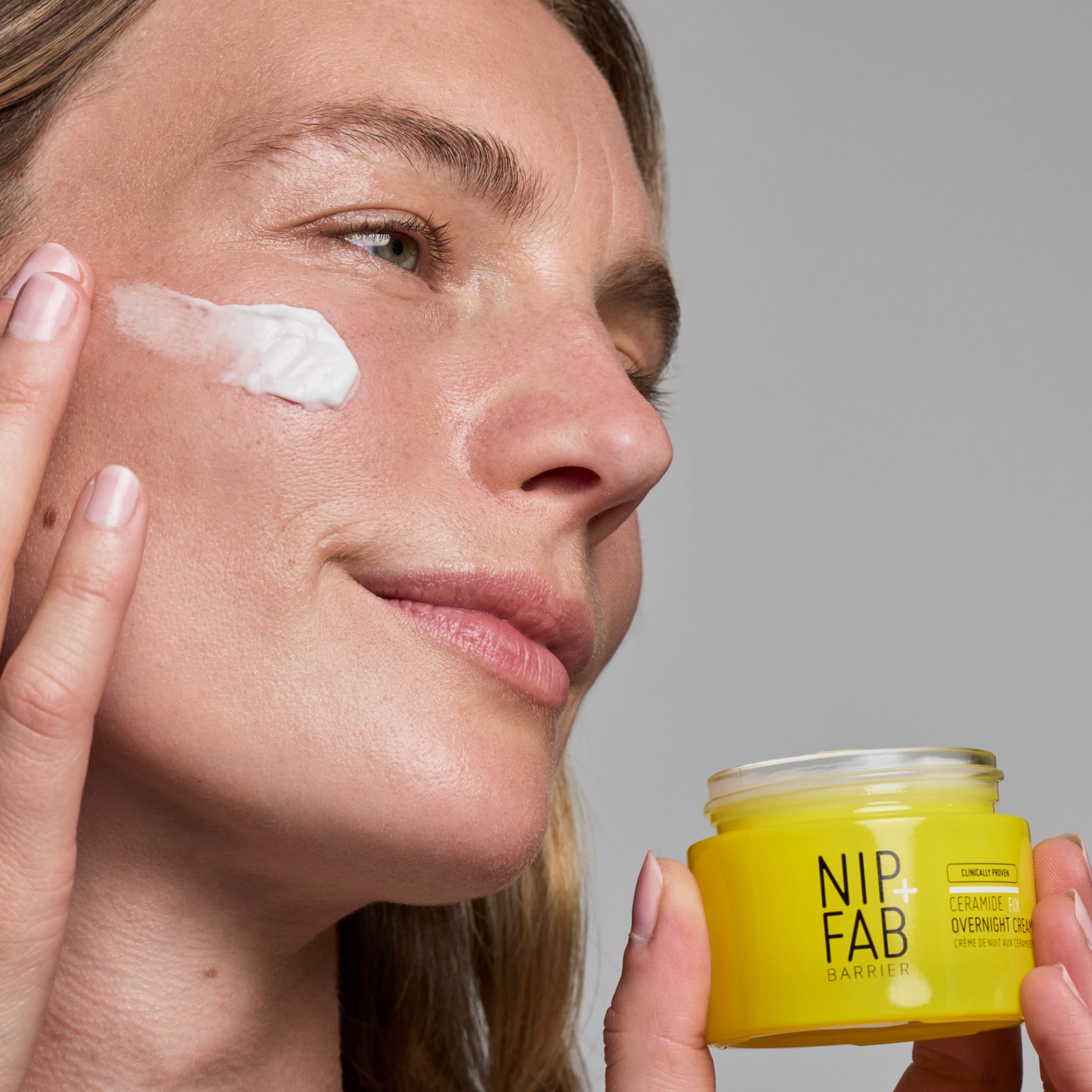 Woman applying cream to her face with a jar of NIP+FAB cream in the foreground.